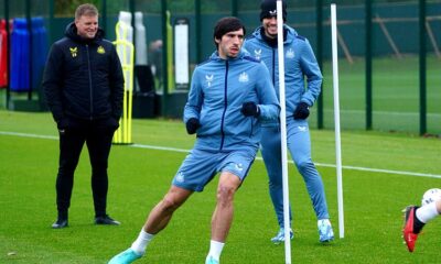 Newcastle United midfielder Sandro Tonali (center) performing agility drills during a training session at the club's Benton base. He is wearing a blue long-sleeved training top and matching shorts. Standing behind him is teammate Bruno Guimarães in similar blue training gear and a black beanie, while manager Eddie Howe watches from the side in a black Castore training jacket. Image used for Sportxparte news.