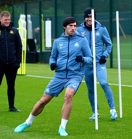 Newcastle United midfielder Sandro Tonali (center) performing agility drills during a training session at the club's Benton base. He is wearing a blue long-sleeved training top and matching shorts. Standing behind him is teammate Bruno Guimarães in similar blue training gear and a black beanie, while manager Eddie Howe watches from the side in a black Castore training jacket. Image used for Sportxparte news.