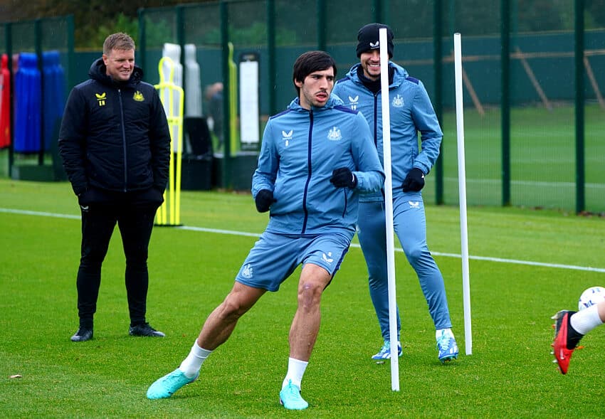 Newcastle United midfielder Sandro Tonali (center) performing agility drills during a training session at the club's Benton base. He is wearing a blue long-sleeved training top and matching shorts. Standing behind him is teammate Bruno Guimarães in similar blue training gear and a black beanie, while manager Eddie Howe watches from the side in a black Castore training jacket. Image used for Sportxparte news.