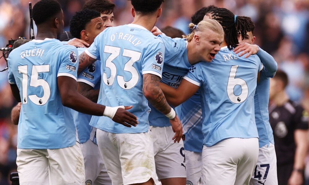 A high-energy photo of several Manchester City football players in their light blue home jerseys, huddled together in a joyous celebration on the pitch. In the center, Erling Haaland is seen with a wide smile, being embraced by teammates including Nico O'Reilly (wearing number 33), Nathan Aké (number 6), and Phil Foden (number 47). The background shows a blurred, crowded Etihad Stadium during their 2-1 Premier League victory against Arsenal. Image used for Sportxparte news.