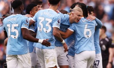 A high-energy photo of several Manchester City football players in their light blue home jerseys, huddled together in a joyous celebration on the pitch. In the center, Erling Haaland is seen with a wide smile, being embraced by teammates including Nico O'Reilly (wearing number 33), Nathan Aké (number 6), and Phil Foden (number 47). The background shows a blurred, crowded Etihad Stadium during their 2-1 Premier League victory against Arsenal. Image used for Sportxparte news.