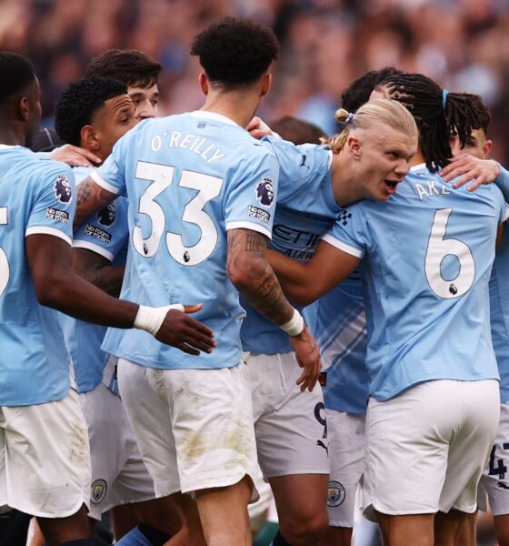 A high-energy photo of several Manchester City football players in their light blue home jerseys, huddled together in a joyous celebration on the pitch. In the center, Erling Haaland is seen with a wide smile, being embraced by teammates including Nico O'Reilly (wearing number 33), Nathan Aké (number 6), and Phil Foden (number 47). The background shows a blurred, crowded Etihad Stadium during their 2-1 Premier League victory against Arsenal. Image used for Sportxparte news.