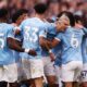 A high-energy photo of several Manchester City football players in their light blue home jerseys, huddled together in a joyous celebration on the pitch. In the center, Erling Haaland is seen with a wide smile, being embraced by teammates including Nico O'Reilly (wearing number 33), Nathan Aké (number 6), and Phil Foden (number 47). The background shows a blurred, crowded Etihad Stadium during their 2-1 Premier League victory against Arsenal. Image used for Sportxparte news.