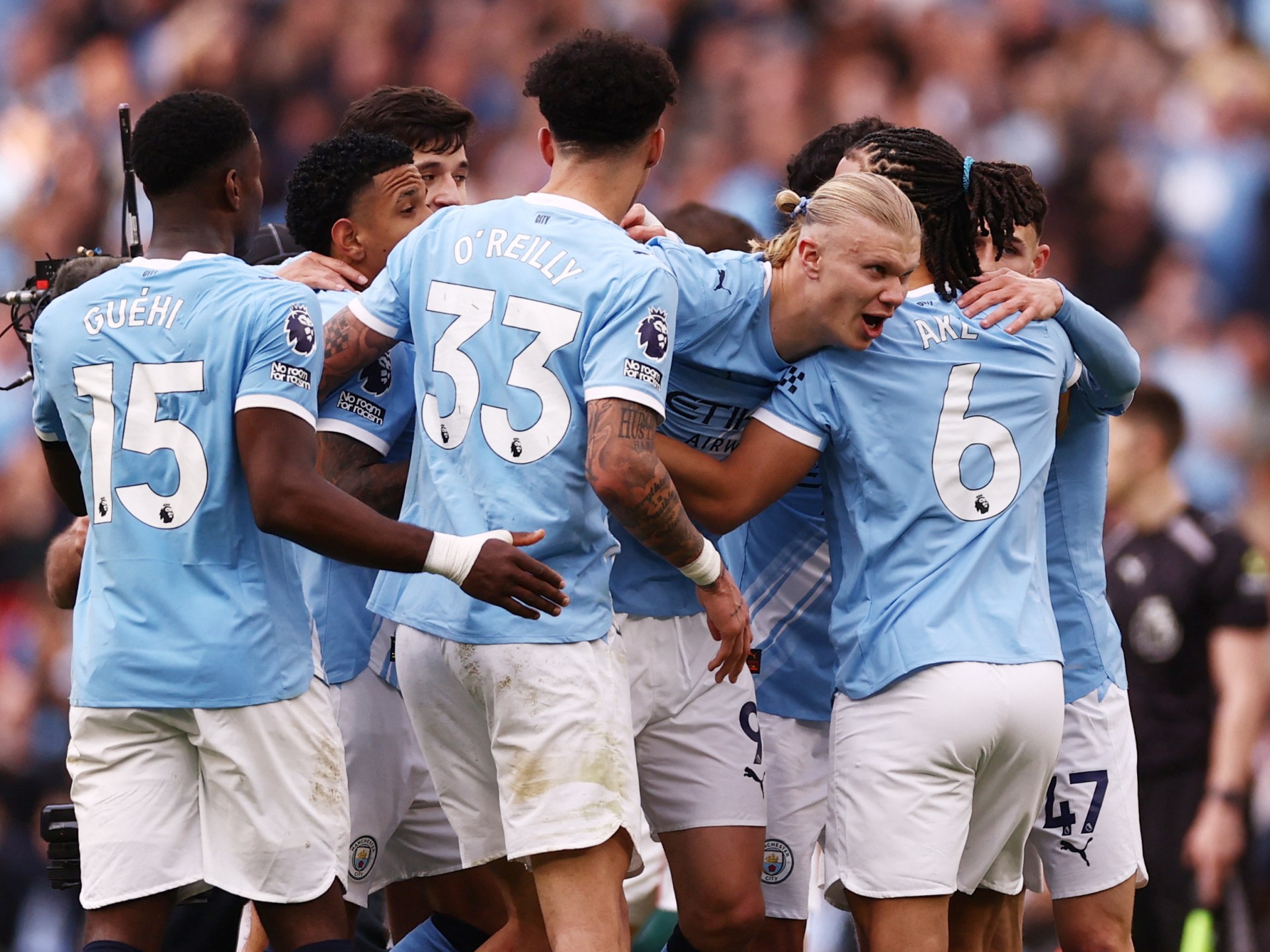 A high-energy photo of several Manchester City football players in their light blue home jerseys, huddled together in a joyous celebration on the pitch. In the center, Erling Haaland is seen with a wide smile, being embraced by teammates including Nico O'Reilly (wearing number 33), Nathan Aké (number 6), and Phil Foden (number 47). The background shows a blurred, crowded Etihad Stadium during their 2-1 Premier League victory against Arsenal. Image used for Sportxparte news.