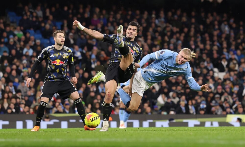Manchester City striker Erling Haaland (right, in light blue) is being challenged by Leeds United defender Pascal Struijk (center, in black/patterned kit). Leeds player James Justin is also visible on the left. Image used for Sportxparte news.