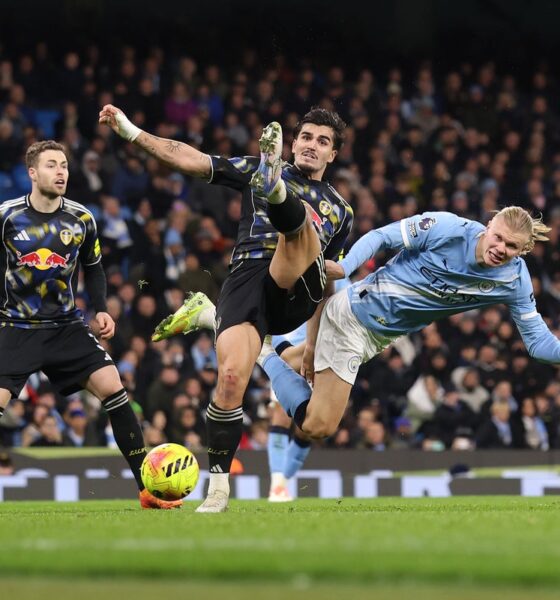 Manchester City striker Erling Haaland (right, in light blue) is being challenged by Leeds United defender Pascal Struijk (center, in black/patterned kit). Leeds player James Justin is also visible on the left. Image used for Sportxparte news.