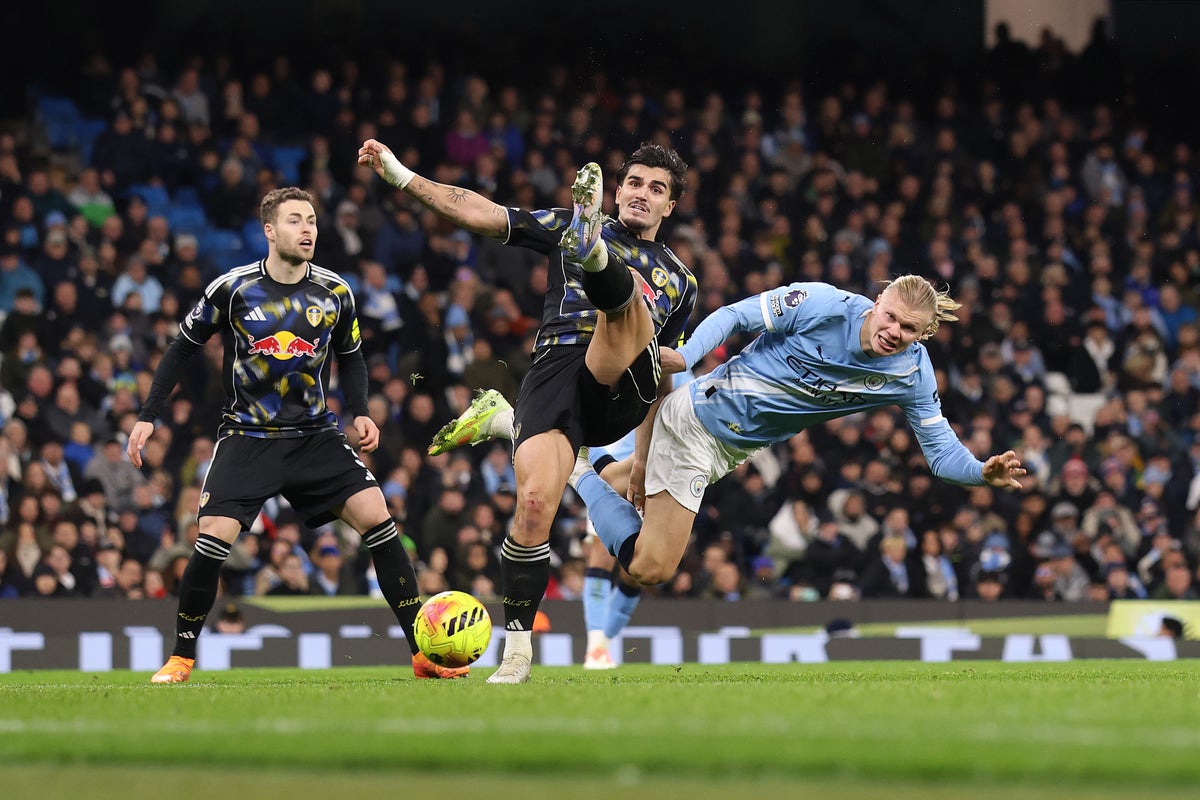 Manchester City striker Erling Haaland (right, in light blue) is being challenged by Leeds United defender Pascal Struijk (center, in black/patterned kit). Leeds player James Justin is also visible on the left. Image used for Sportxparte news.