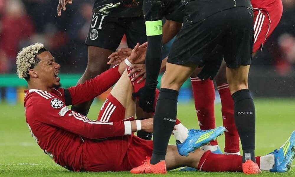 A close-up shot of Hugo Ekitike in a red Liverpool kit, sitting on the grass at Anfield and clutching his lower right leg in agony. He is surrounded by concerned players from both teams, including a PSG player in a black kit leaning over him. The image captures the moment the French striker was forced off on a stretcher during the 27th minute of the second-leg clash on April 14, 2026. Image used for Sportxparte news.