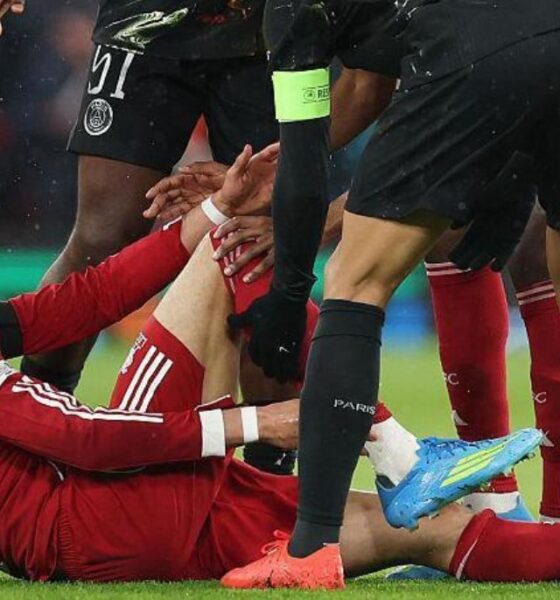 A close-up shot of Hugo Ekitike in a red Liverpool kit, sitting on the grass at Anfield and clutching his lower right leg in agony. He is surrounded by concerned players from both teams, including a PSG player in a black kit leaning over him. The image captures the moment the French striker was forced off on a stretcher during the 27th minute of the second-leg clash on April 14, 2026. Image used for Sportxparte news.