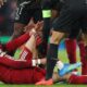 A close-up shot of Hugo Ekitike in a red Liverpool kit, sitting on the grass at Anfield and clutching his lower right leg in agony. He is surrounded by concerned players from both teams, including a PSG player in a black kit leaning over him. The image captures the moment the French striker was forced off on a stretcher during the 27th minute of the second-leg clash on April 14, 2026. Image used for Sportxparte news.