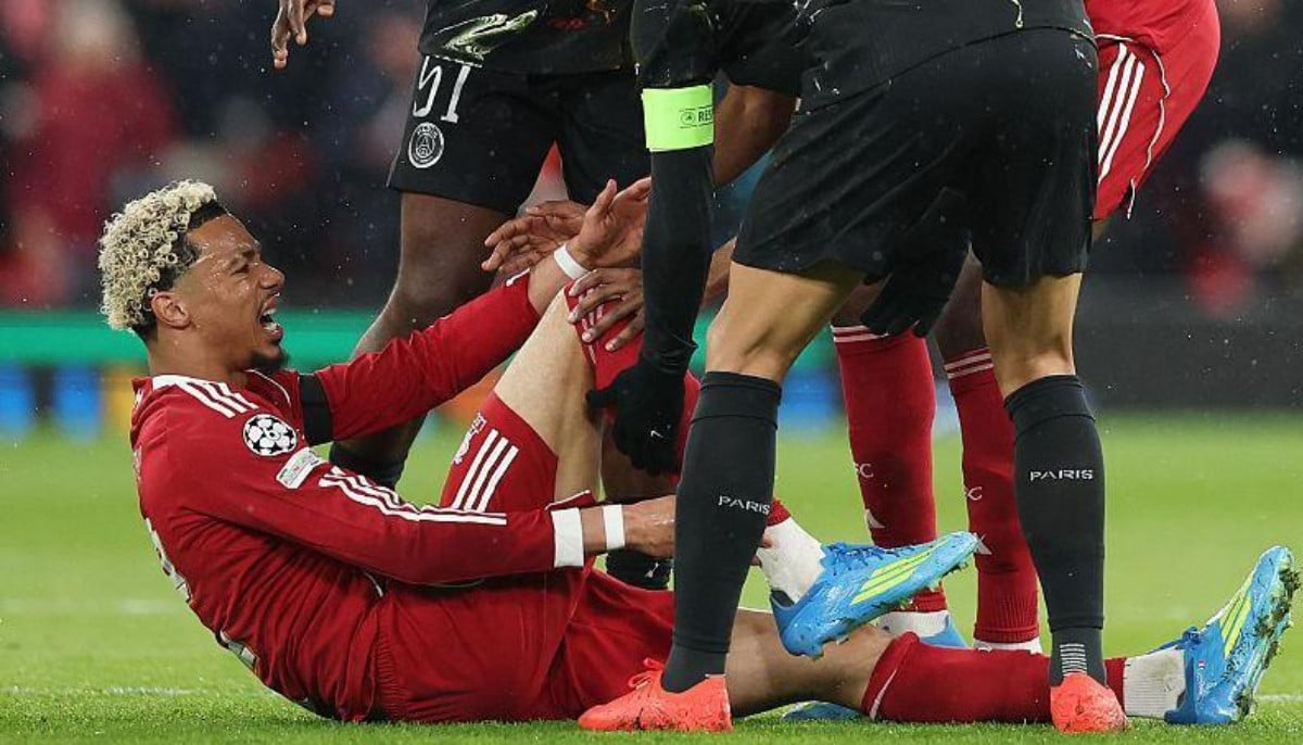 A close-up shot of Hugo Ekitike in a red Liverpool kit, sitting on the grass at Anfield and clutching his lower right leg in agony. He is surrounded by concerned players from both teams, including a PSG player in a black kit leaning over him. The image captures the moment the French striker was forced off on a stretcher during the 27th minute of the second-leg clash on April 14, 2026. Image used for Sportxparte news.
