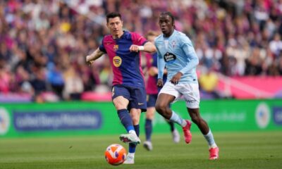 Robert Lewandowski, in a dark blue and red FC Barcelona kit, shields the ball and prepares to strike while being closely pursued by Ilaix Moriba in a light blue Celta Vigo jersey during a La Liga match at a crowded stadium. Image used for Sportxparte news.
