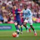 Robert Lewandowski, in a dark blue and red FC Barcelona kit, shields the ball and prepares to strike while being closely pursued by Ilaix Moriba in a light blue Celta Vigo jersey during a La Liga match at a crowded stadium. Image used for Sportxparte news.