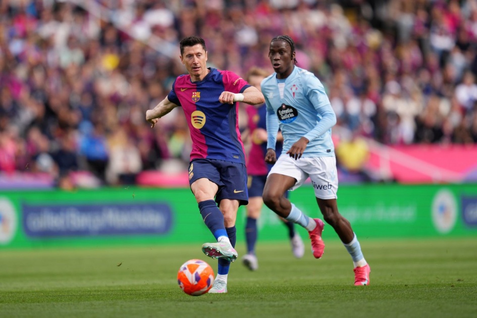 Robert Lewandowski, in a dark blue and red FC Barcelona kit, shields the ball and prepares to strike while being closely pursued by Ilaix Moriba in a light blue Celta Vigo jersey during a La Liga match at a crowded stadium. Image used for Sportxparte news.
