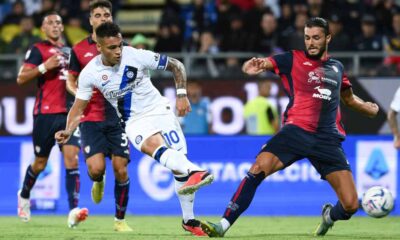 Inter Milan forward Lautaro Martínez, wearing a white away kit with the number 10, strikes a low shot toward the goal while Cagliari defender Alberto Dossena slides in to block during a Serie A football match at the Sardegna Arena. Image used for Sportxparte news.