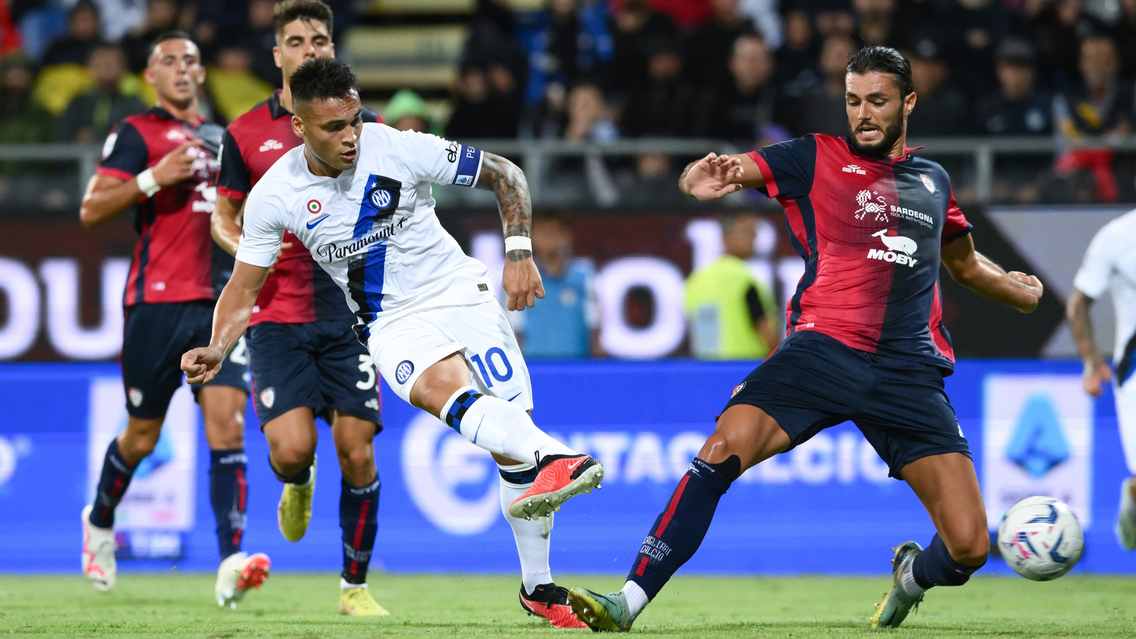 Inter Milan forward Lautaro Martínez, wearing a white away kit with the number 10, strikes a low shot toward the goal while Cagliari defender Alberto Dossena slides in to block during a Serie A football match at the Sardegna Arena. Image used for Sportxparte news.