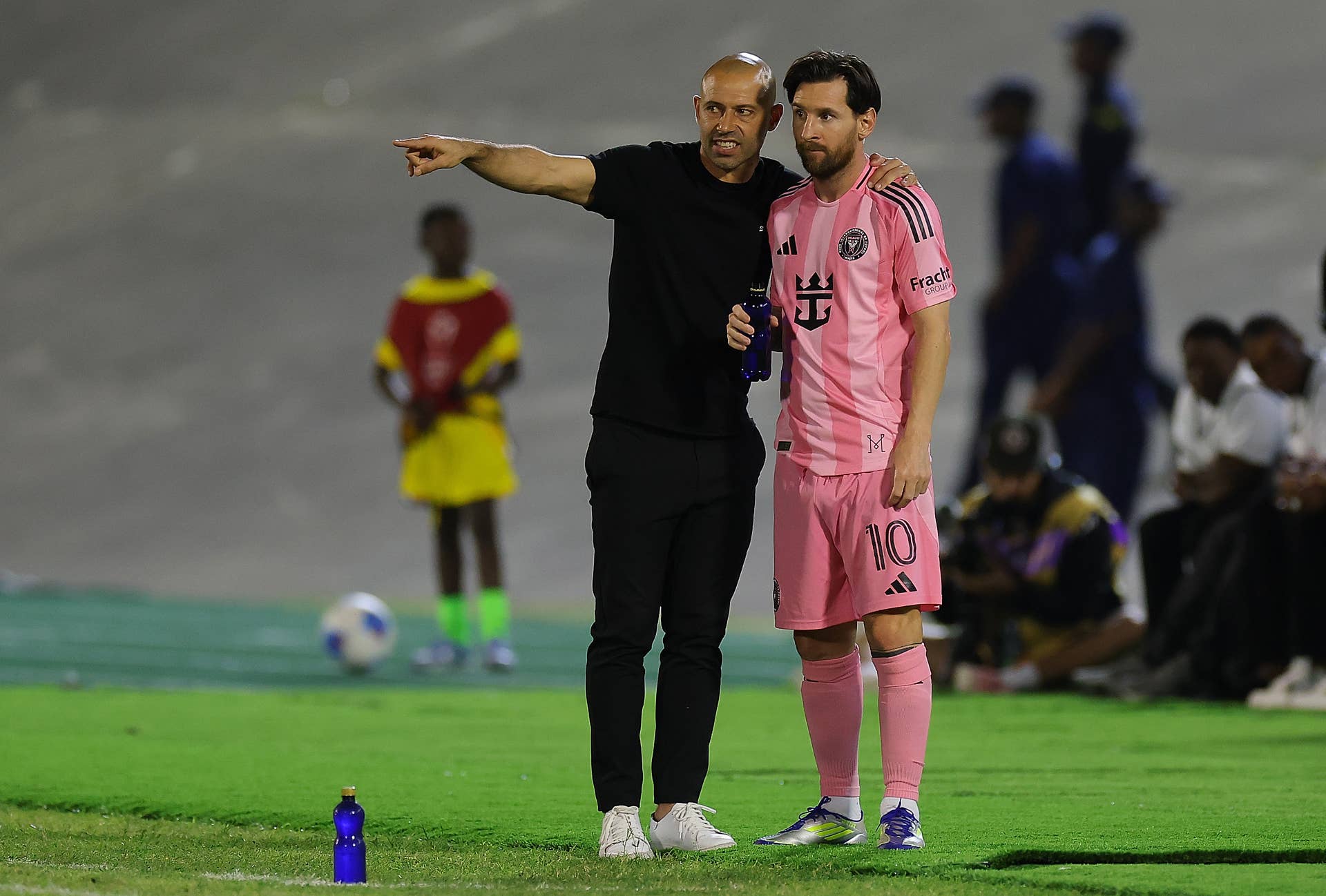 Lionel Messi and coach Javier Mascherano on the pitch in the Inter Miami 24/25 Home Authentic Jersey. Messi wears the pink "Euforia" kit with his iconic number 10, standing next to Mascherano in all-black sideline gear during an Inter Miami CF match. Image used for Sportxparte news.