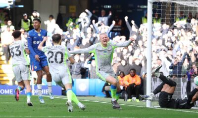 Manchester City's Nico O'Reilly celebrates with high energy after scoring the opening goal during the Premier League match against Chelsea at Stamford Bridge on April 12, 2026. The young midfielder is shown in City's grey and yellow away kit, with the Chelsea goal and reacting players visible in the background. Image used for Sportxparte news