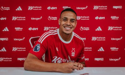 Brazilian defender Murillo wearing the red Nottingham Forest home kit, smiles and holds a pen over contract documents against a club-branded sponsor backdrop. Image used by Sportxparte for newsd