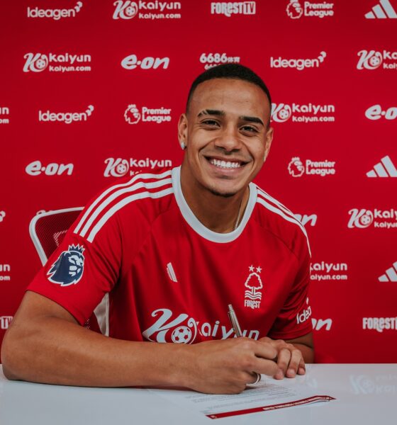 Brazilian defender Murillo wearing the red Nottingham Forest home kit, smiles and holds a pen over contract documents against a club-branded sponsor backdrop. Image used by Sportxparte for newsd