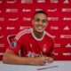 Brazilian defender Murillo wearing the red Nottingham Forest home kit, smiles and holds a pen over contract documents against a club-branded sponsor backdrop. Image used by Sportxparte for newsd