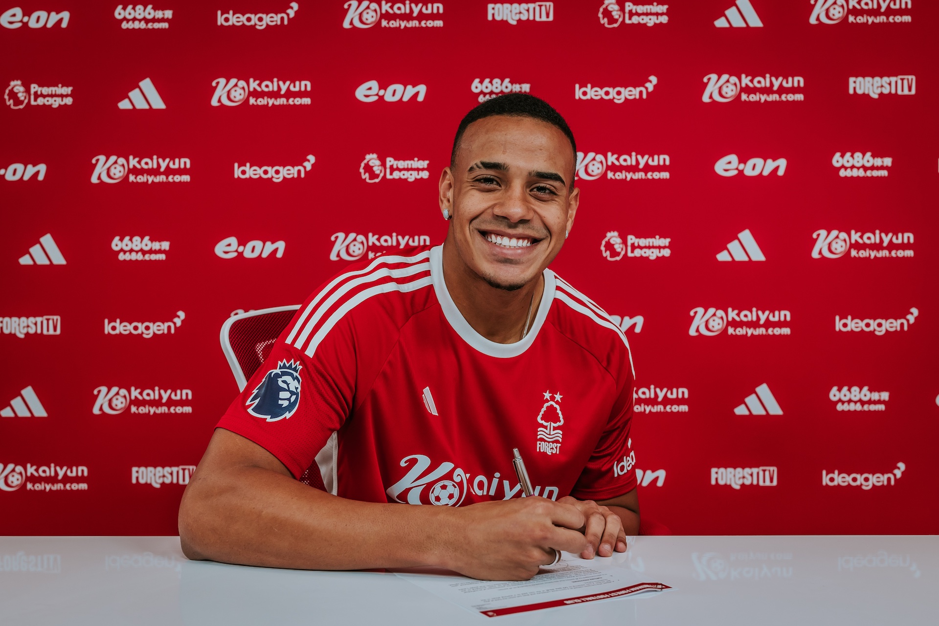 Brazilian defender Murillo wearing the red Nottingham Forest home kit, smiles and holds a pen over contract documents against a club-branded sponsor backdrop. Image used by Sportxparte for newsd