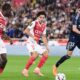 The image captures a Ligue 1 football match between AS Monaco and Paris FC. The featured players include Maghnes Akliouche (center, wearing #11) and George Ilenikhena (left, wearing #28) for AS Monaco, and Pierre Lees-Melou (right, wearing #33) for Paris FC. Image used for Sportxparte news
