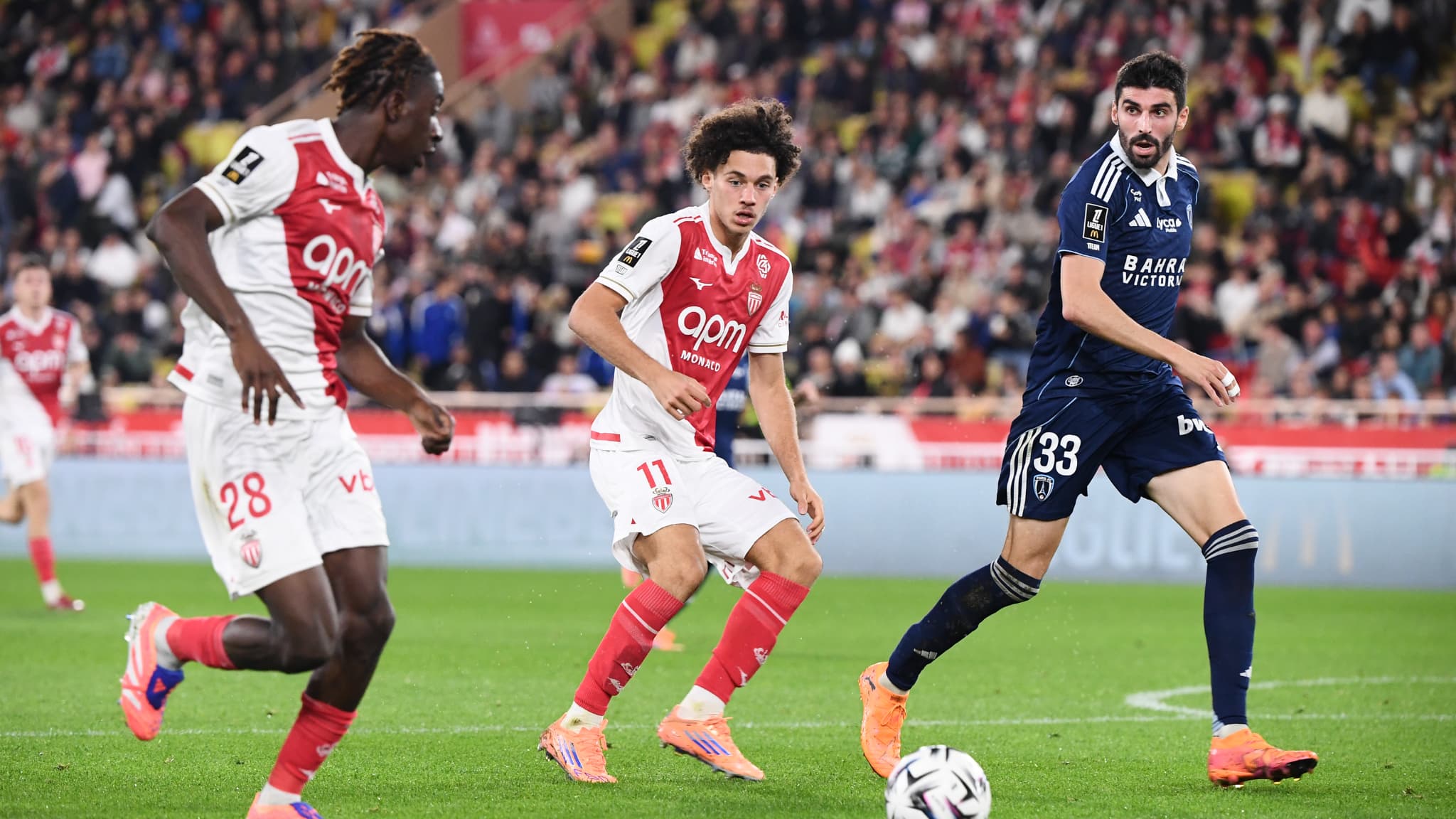 The image captures a Ligue 1 football match between AS Monaco and Paris FC. The featured players include Maghnes Akliouche (center, wearing #11) and George Ilenikhena (left, wearing #28) for AS Monaco, and Pierre Lees-Melou (right, wearing #33) for Paris FC. Image used for Sportxparte news