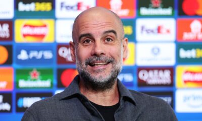A close-up of a smiling Pep Guardiola, the manager of Manchester City, with a salt-and-pepper beard. He is wearing a dark grey, textured button-down shirt over a black t-shirt, standing in front of a blue press conference backdrop featuring various corporate logos. Image used for Sportxparte news.