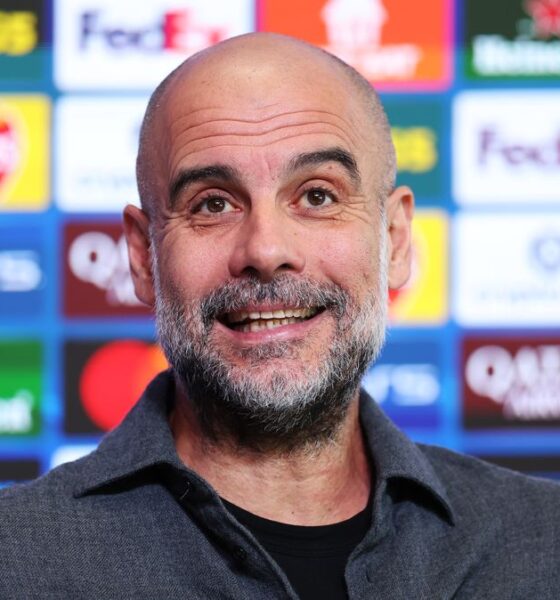 A close-up of a smiling Pep Guardiola, the manager of Manchester City, with a salt-and-pepper beard. He is wearing a dark grey, textured button-down shirt over a black t-shirt, standing in front of a blue press conference backdrop featuring various corporate logos. Image used for Sportxparte news.