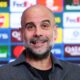 A close-up of a smiling Pep Guardiola, the manager of Manchester City, with a salt-and-pepper beard. He is wearing a dark grey, textured button-down shirt over a black t-shirt, standing in front of a blue press conference backdrop featuring various corporate logos. Image used for Sportxparte news.
