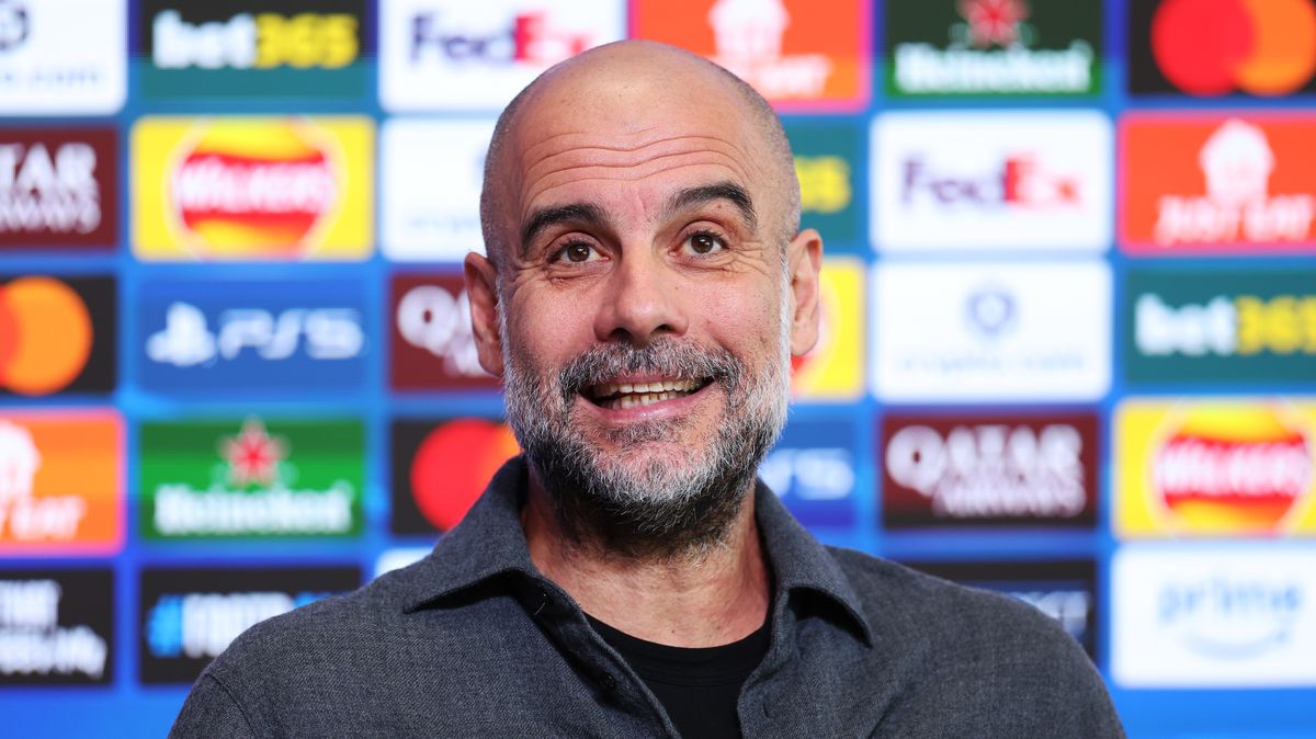 A close-up of a smiling Pep Guardiola, the manager of Manchester City, with a salt-and-pepper beard. He is wearing a dark grey, textured button-down shirt over a black t-shirt, standing in front of a blue press conference backdrop featuring various corporate logos. Image used for Sportxparte news.