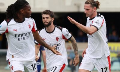 Three AC Milan players, Rafael Leão (#10), Christian Pulisic (#11), and Adrien Rabiot (#12), celebrate on the pitch during a Serie A match against Hellas Verona. They are wearing the club's white 2025-26 PUMA away kit. Rafael Leão is on the left, looking toward his teammates; Christian Pulisic is in the center; and Adrien Rabiot is on the right with his arm raised in celebration. Image used for Sportxparte news.
