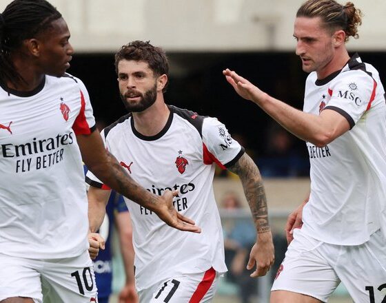 Three AC Milan players, Rafael Leão (#10), Christian Pulisic (#11), and Adrien Rabiot (#12), celebrate on the pitch during a Serie A match against Hellas Verona. They are wearing the club's white 2025-26 PUMA away kit. Rafael Leão is on the left, looking toward his teammates; Christian Pulisic is in the center; and Adrien Rabiot is on the right with his arm raised in celebration. Image used for Sportxparte news.