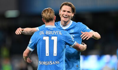 Kevin De Bruyne and Scott McTominay celebrate together during a soccer match. De Bruyne, with his back to the camera, wears a light blue SSC Napoli jersey with his name and the number 11 prominently displayed. McTominay, facing the camera with a joyful expression, has his arm around De Bruyne's shoulder. Image used for Sportxparte news roundup.