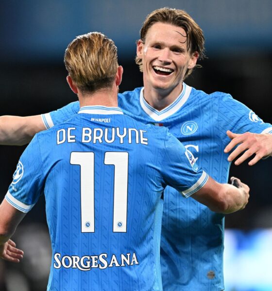 Kevin De Bruyne and Scott McTominay celebrate together during a soccer match. De Bruyne, with his back to the camera, wears a light blue SSC Napoli jersey with his name and the number 11 prominently displayed. McTominay, facing the camera with a joyful expression, has his arm around De Bruyne's shoulder. Image used for Sportxparte news roundup.