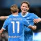 Kevin De Bruyne and Scott McTominay celebrate together during a soccer match. De Bruyne, with his back to the camera, wears a light blue SSC Napoli jersey with his name and the number 11 prominently displayed. McTominay, facing the camera with a joyful expression, has his arm around De Bruyne's shoulder. Image used for Sportxparte news roundup.