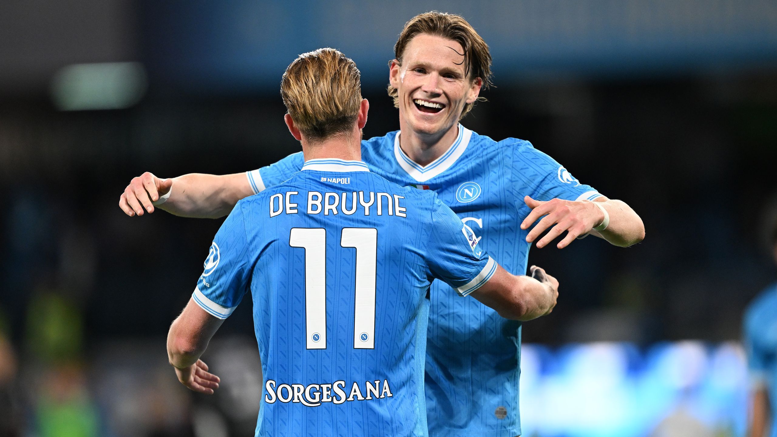 Kevin De Bruyne and Scott McTominay celebrate together during a soccer match. De Bruyne, with his back to the camera, wears a light blue SSC Napoli jersey with his name and the number 11 prominently displayed. McTominay, facing the camera with a joyful expression, has his arm around De Bruyne's shoulder. Image used for Sportxparte news roundup.