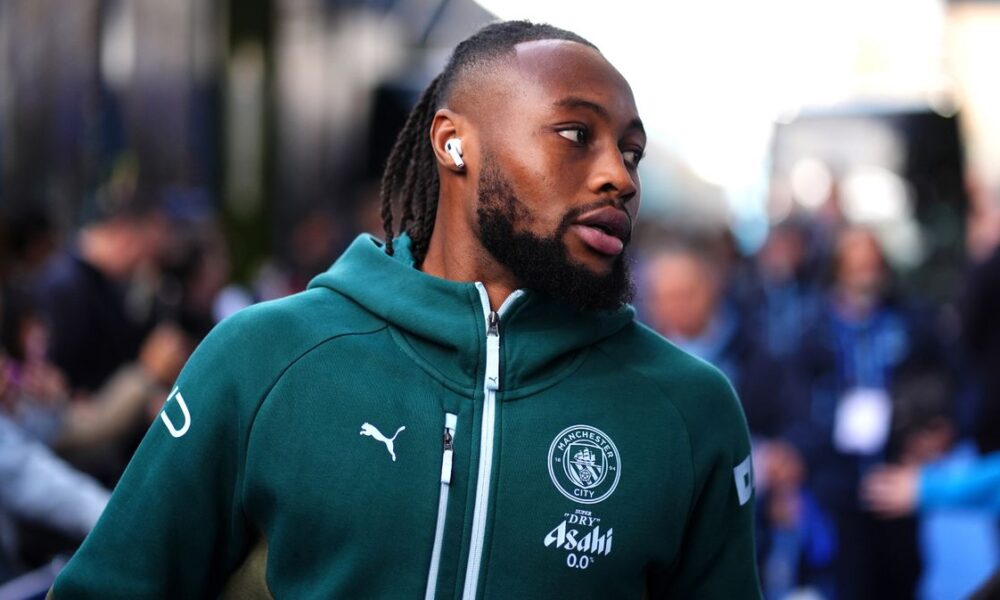 A close-up of professional footballer Antoine Semenyo wearing a teal Manchester City training hoodie. He is looking off-camera to his right with a focused expression, and a white wireless earbud is visible in his ear. Image used for Sportxparte news.