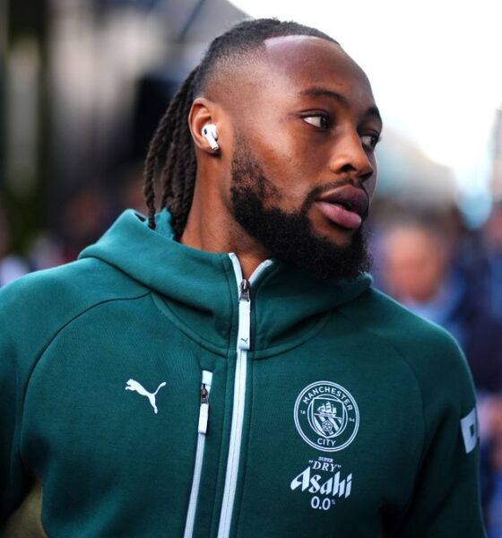 A close-up of professional footballer Antoine Semenyo wearing a teal Manchester City training hoodie. He is looking off-camera to his right with a focused expression, and a white wireless earbud is visible in his ear. Image used for Sportxparte news.