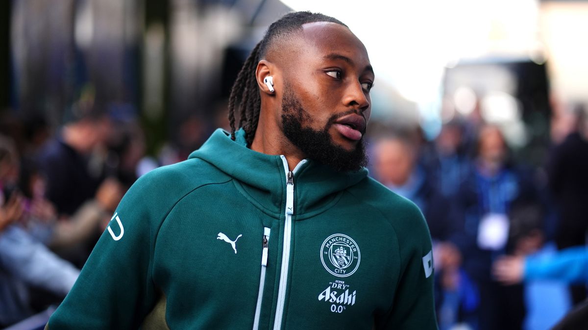 A close-up of professional footballer Antoine Semenyo wearing a teal Manchester City training hoodie. He is looking off-camera to his right with a focused expression, and a white wireless earbud is visible in his ear. Image used for Sportxparte news.