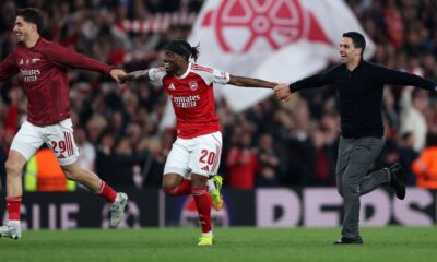 Arsenal manager, Mikel Arteta, and two players, Noni Madueke and Kai Havertz, are running across a football pitch with joyous expressions, celebrating a victory. Arteta is in a dark blue sweater and grey trousers, while the players wear their red and white team kits. Image used for Sportxparte news.