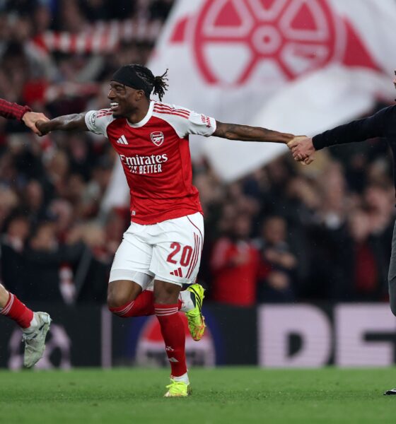 Arsenal manager, Mikel Arteta, and two players, Noni Madueke and Kai Havertz, are running across a football pitch with joyous expressions, celebrating a victory. Arteta is in a dark blue sweater and grey trousers, while the players wear their red and white team kits. Image used for Sportxparte news.