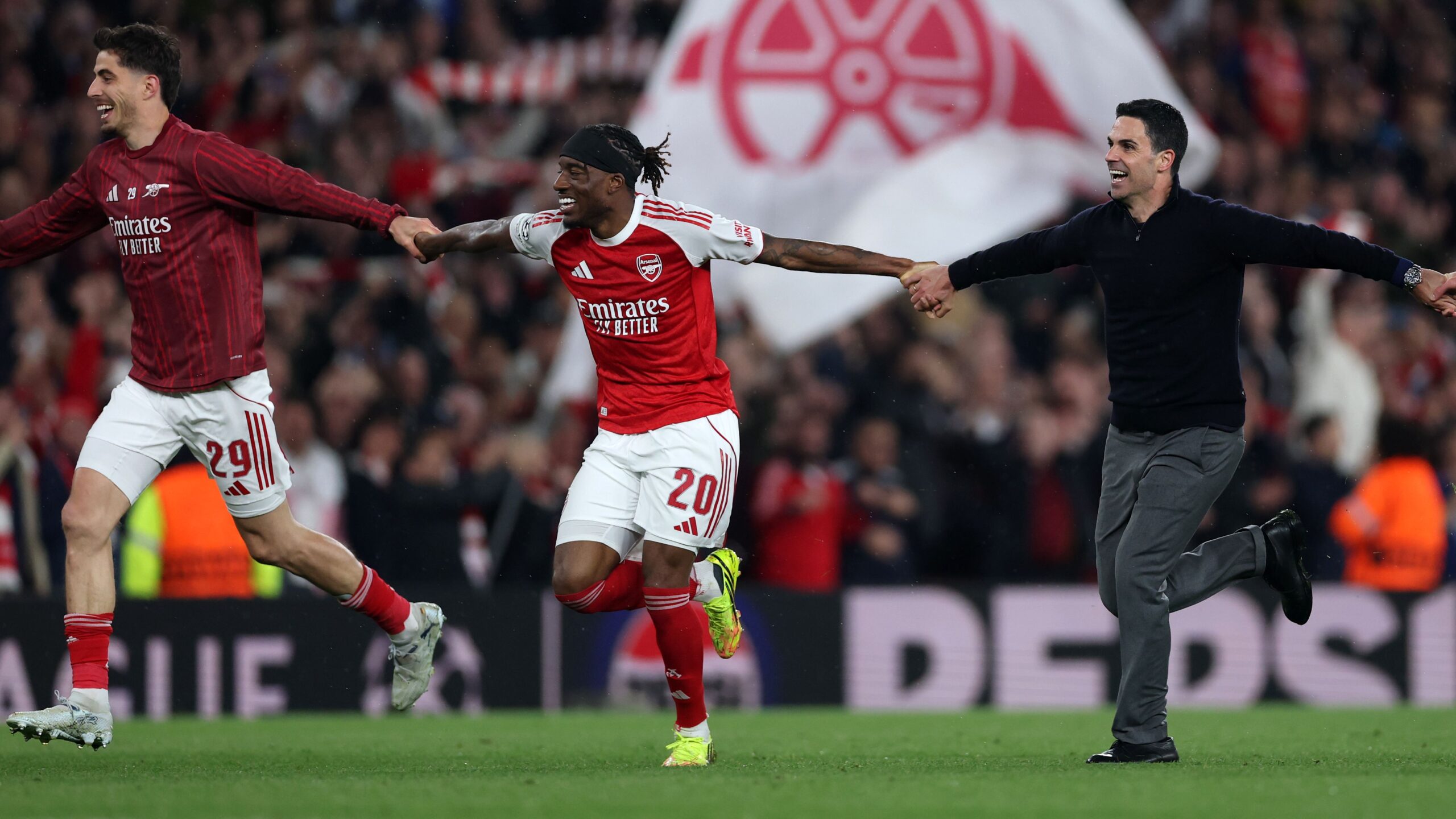 Arsenal manager, Mikel Arteta, and two players, Noni Madueke and Kai Havertz, are running across a football pitch with joyous expressions, celebrating a victory. Arteta is in a dark blue sweater and grey trousers, while the players wear their red and white team kits. Image used for Sportxparte news.