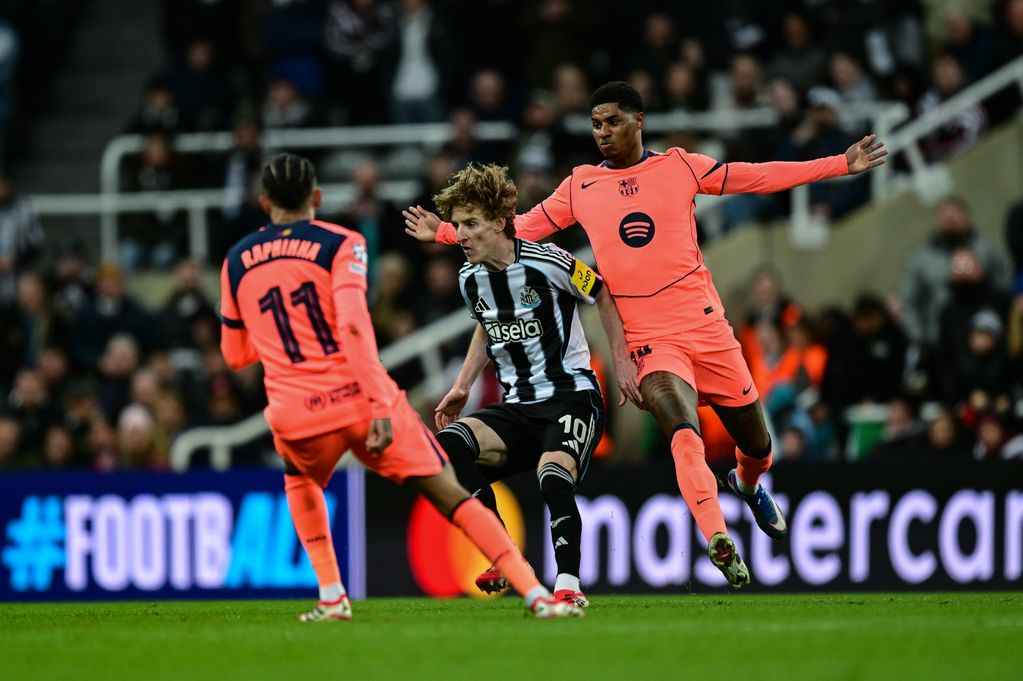 An action shot from a UEFA Champions League match at St James' Park shows FC Barcelona forward Marcus Rashford in a coral-orange kit battling for possession against Newcastle United's Anthony Gordon, who is wearing a black-and-white striped jersey, while Barcelona's Raphinha approaches the play from behind. Image used for Sportxparte news.