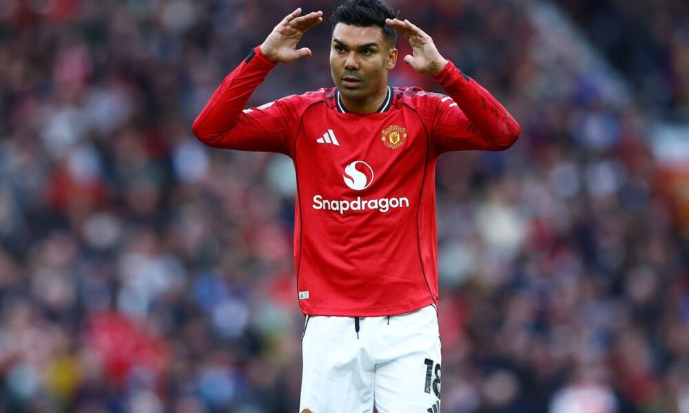 Manchester United football player Casemiro on the pitch. He is wearing the red 2024/2025 home kit with white shorts. He has both hands raised to the sides of his head, looking forward with a focused expression. Image used for Sportxparte news.