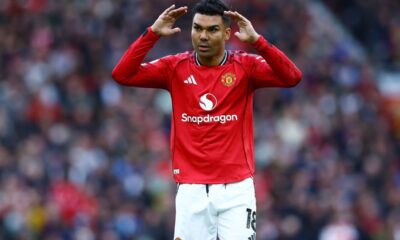 Manchester United football player Casemiro on the pitch. He is wearing the red 2024/2025 home kit with white shorts. He has both hands raised to the sides of his head, looking forward with a focused expression. Image used for Sportxparte news.