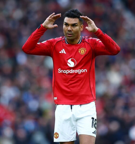 Manchester United football player Casemiro on the pitch. He is wearing the red 2024/2025 home kit with white shorts. He has both hands raised to the sides of his head, looking forward with a focused expression. Image used for Sportxparte news.