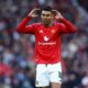 Manchester United football player Casemiro on the pitch. He is wearing the red 2024/2025 home kit with white shorts. He has both hands raised to the sides of his head, looking forward with a focused expression. Image used for Sportxparte news.