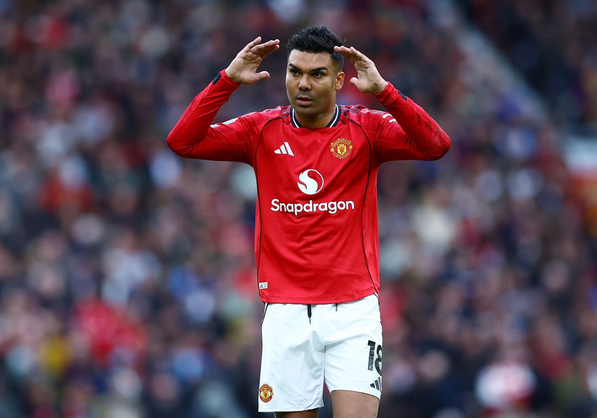Manchester United football player Casemiro on the pitch. He is wearing the red 2024/2025 home kit with white shorts. He has both hands raised to the sides of his head, looking forward with a focused expression. Image used for Sportxparte news.
